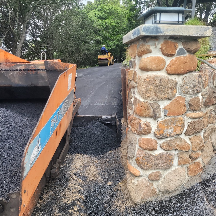 Asphalt paving equipment working beside a stone driveway entrance